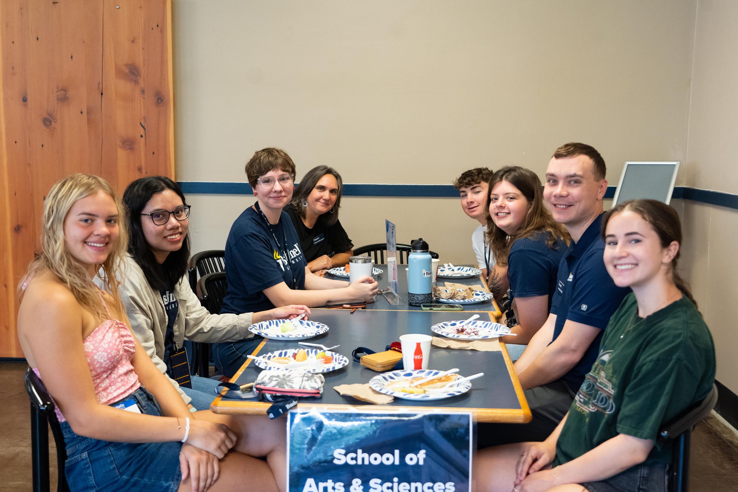 A group of students and professors smile at a cafeteria table during breakfast.