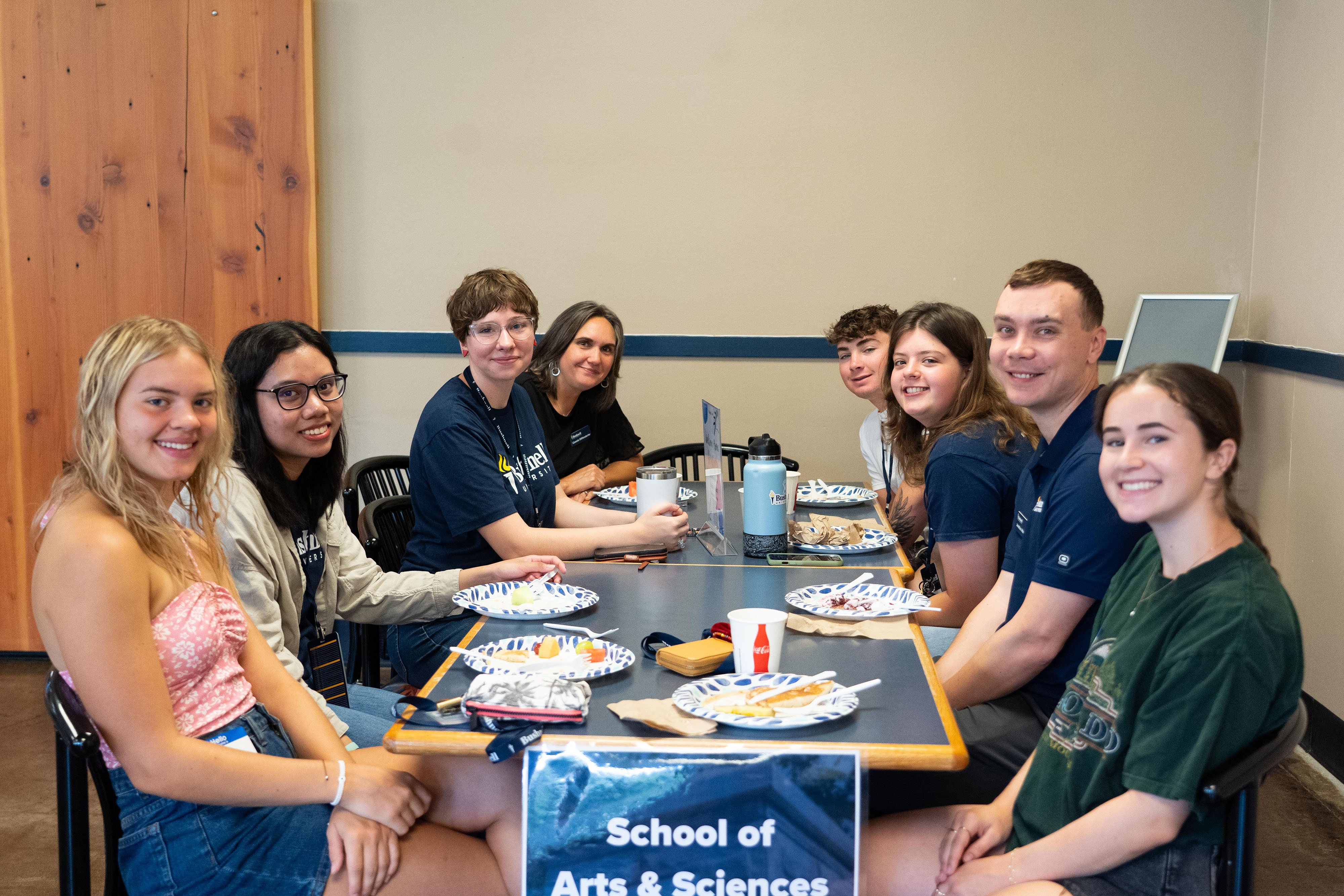 A group of students and professors smile at a cafeteria table during breakfast.