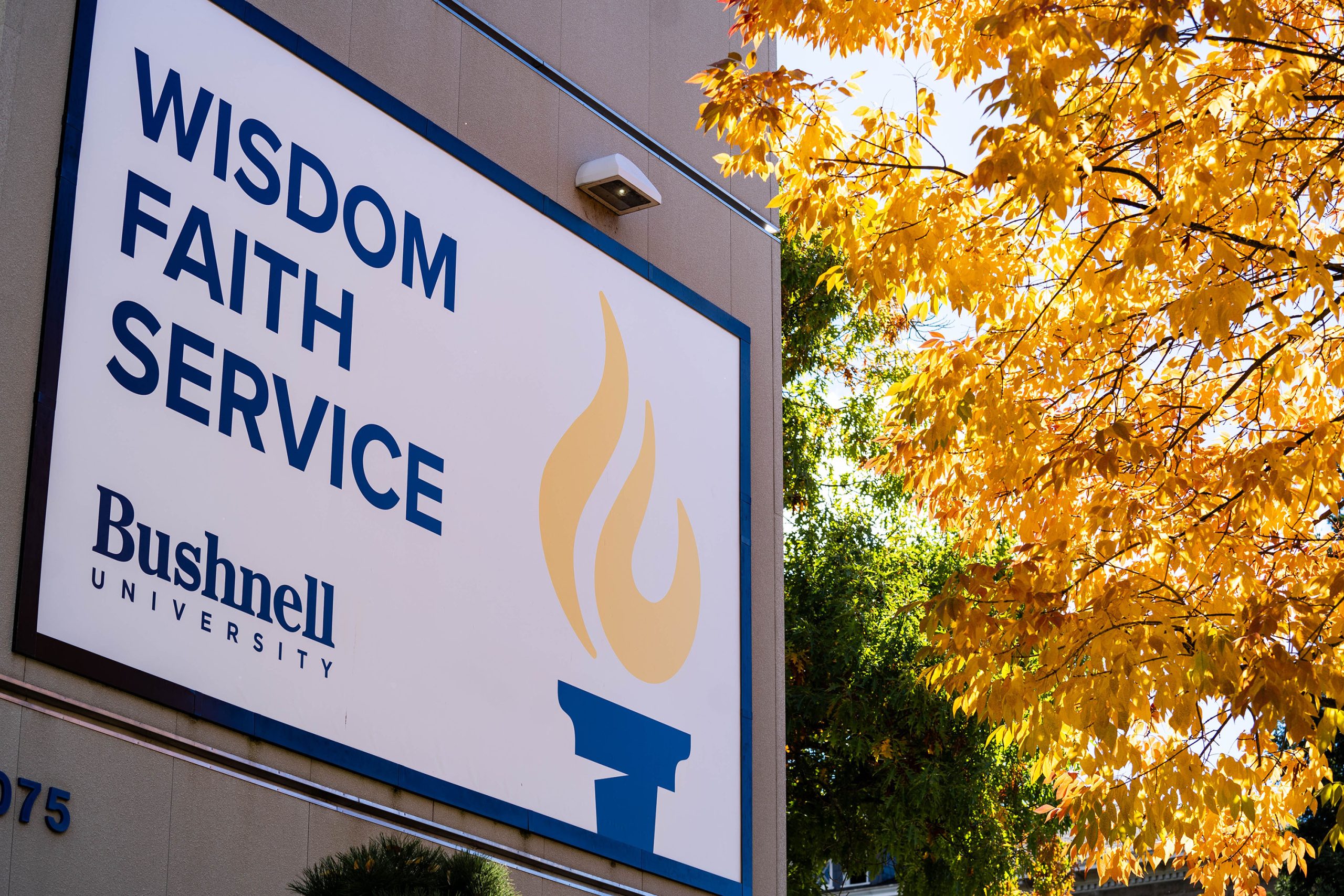 A banner on a campus building with "wisdom, faith, service" in bold and Beacon logo next to a golden leaved tree in the fall.