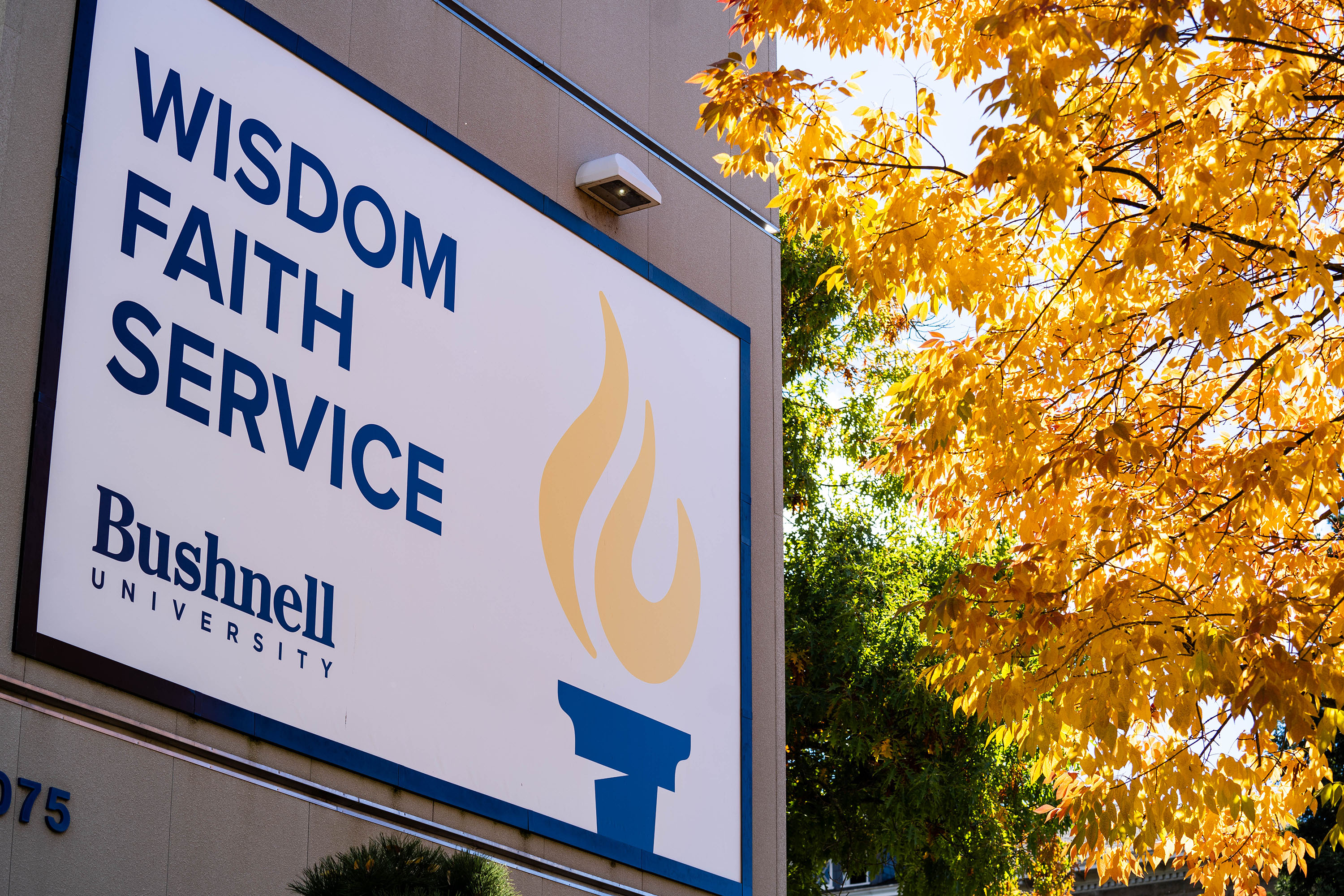 A banner on a campus building with "wisdom, faith, service" in bold and Beacon logo next to a golden leaved tree in the fall.