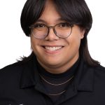Portrait of a smiling person with straight dark hair and glasses wearing a black Bushnell polo shirt against a white background.