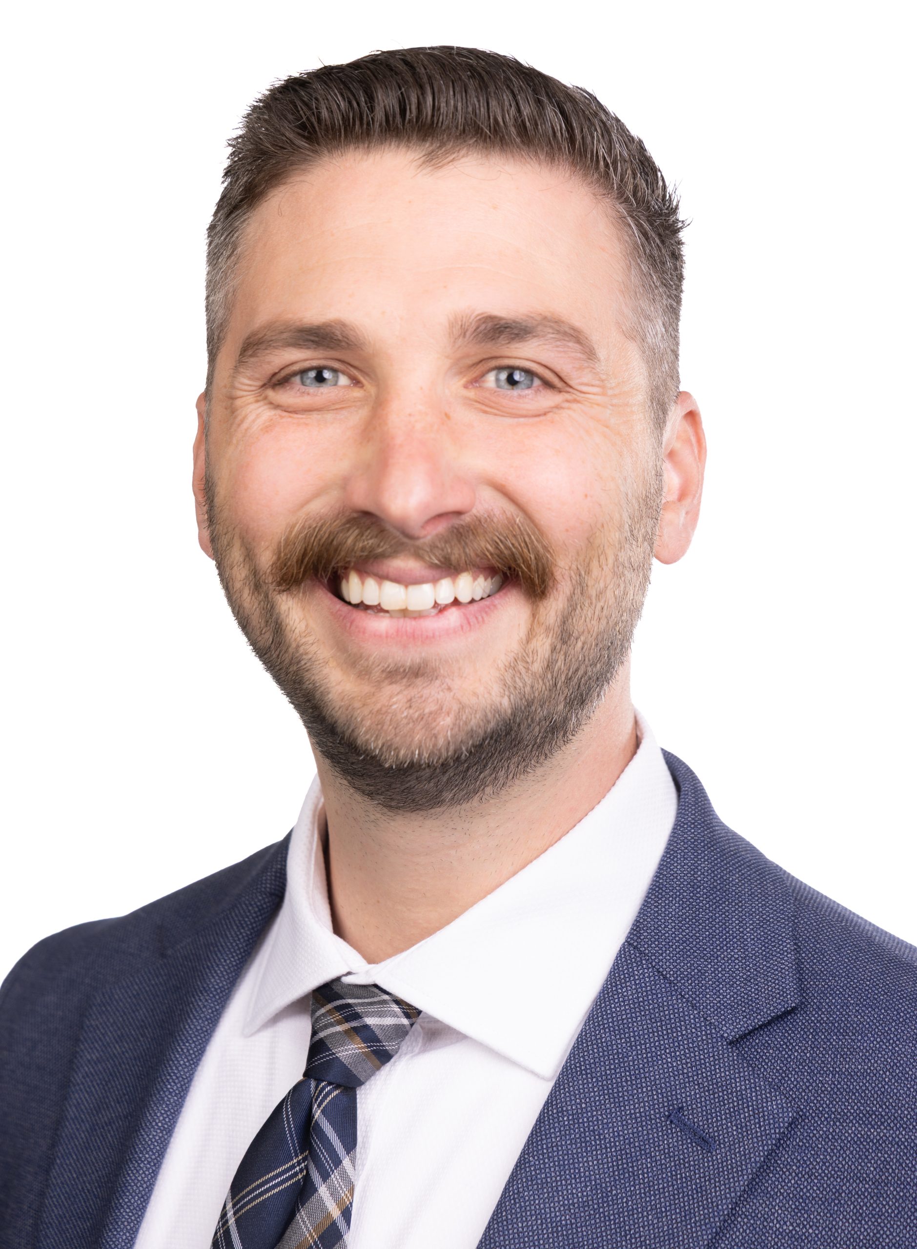 Portrait of a smiling man with short brown hair and a trimmed beard wearing a blue suit and patterned tie against a white background.