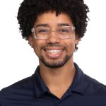 Portrait of a smiling man with curly dark hair and glasses wearing a navy Bushnell University polo shirt against a white background.
