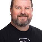 Portrait of a smiling man with short brown hair and a full beard wearing a black Bushnell shirt against a white background.