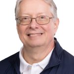 Portrait of a smiling older man with gray hair and glasses wearing a navy Bushnell jacket over a white polo shirt against a white background.