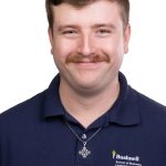 Portrait of a smiling man with short brown hair and a mustache wearing a navy Bushnell School of Business, Leadership, & Technology polo shirt and a cross necklace against a white background.