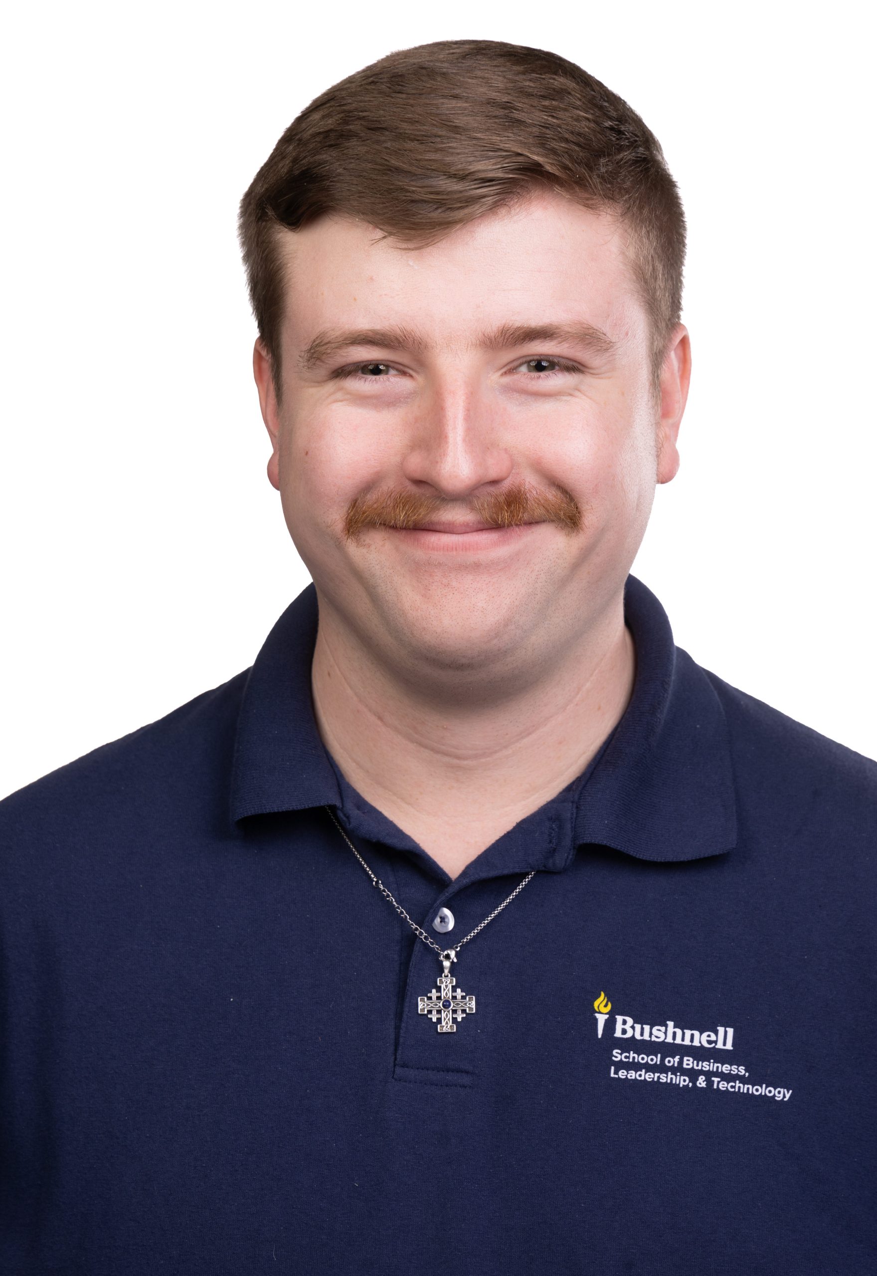 Portrait of a smiling man with short brown hair and a mustache wearing a navy Bushnell School of Business, Leadership, & Technology polo shirt and a cross necklace against a white background.
