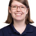 Portrait of a smiling woman with shoulder-length brown hair and glasses wearing a navy Bushnell University polo shirt against a white background.