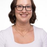 Portrait of a smiling woman with short wavy brown hair and black-framed glasses wearing a white top and delicate necklace against a white background.