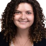 Portrait of a smiling woman with long curly brown hair wearing a navy Bushnell cardigan over a white top against a white background.