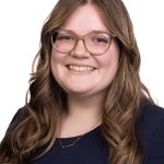 Portrait of a smiling woman with long wavy brown hair and glasses wearing a navy top against a white background.