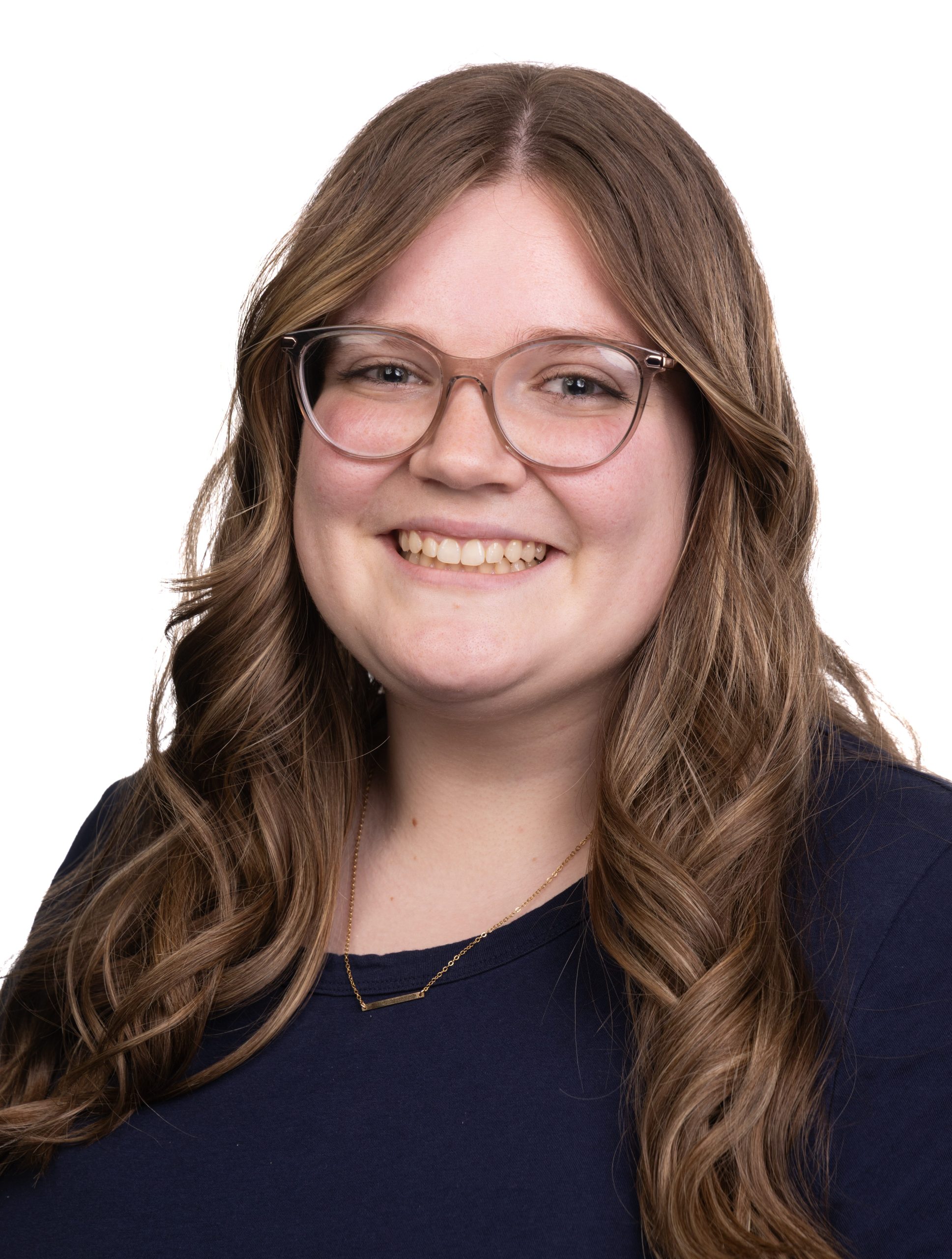 Portrait of a smiling woman with long wavy brown hair and glasses wearing a navy top against a white background.