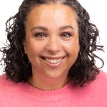 Portrait of a smiling woman with curly dark hair wearing a pink sweater against a white background.