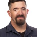 Portrait of a man with short dark brown hair and a full beard wearing a navy Bushnell University collared shirt against a white background.