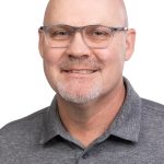 Portrait of a smiling bald man with glasses and a short gray beard wearing a gray Bushnell polo shirt against a white background.