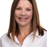 Portrait of a smiling woman with straight shoulder-length brown hair wearing a white collared shirt against a white background.