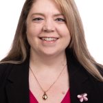 Portrait of a smiling woman with long light brown hair wearing a black blazer over a bright pink top against a white background.