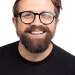 Portrait of a smiling man with short brown hair, a full brown beard, and black-framed glasses wearing a black crew-neck shirt against a white background.