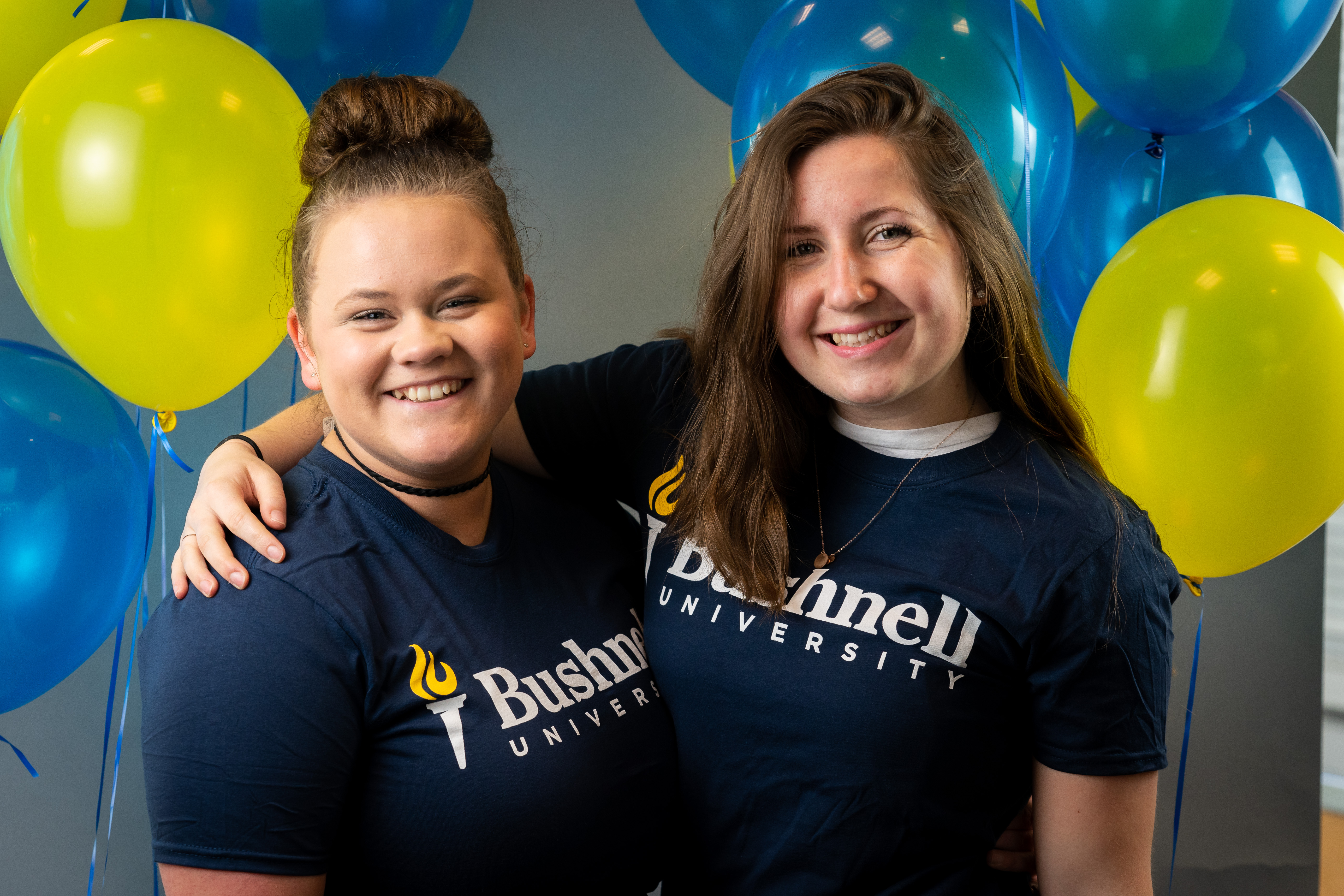 Two Bushnell University students smiling together in front of blue and gold balloons.