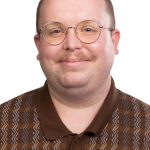Portrait of a man with round glasses and a mustache wearing a brown patterned polo against a white background.