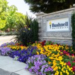 Bushnell University entrance sign surrounded by colorful flowers and green landscaping.