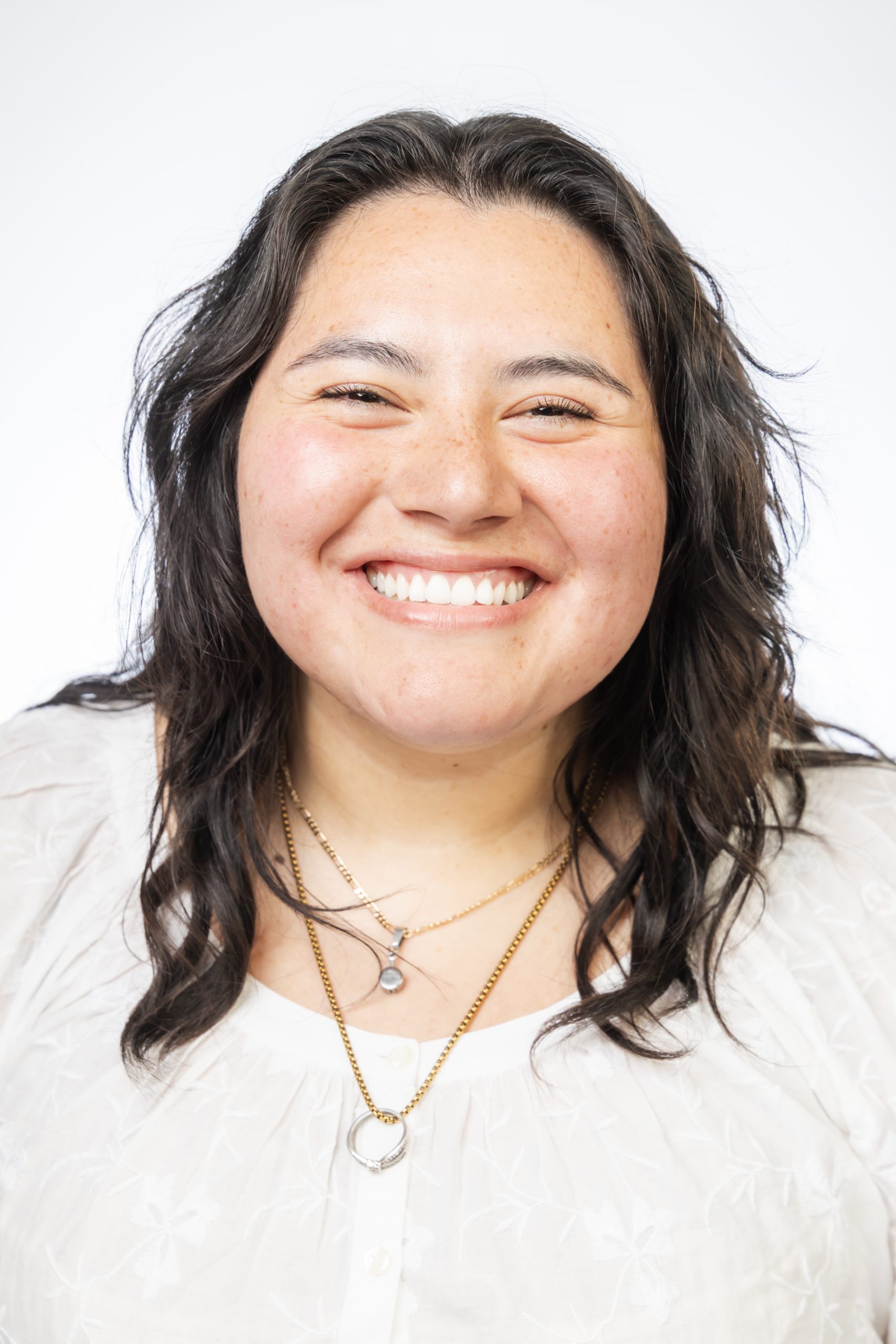 Portrait of a smiling woman with dark hair wearing a white blouse and layered necklaces against a white background.