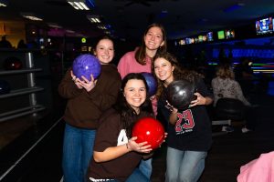 Four students hold bowling balls at a bowling alley during a Beacon Night off-campus event.