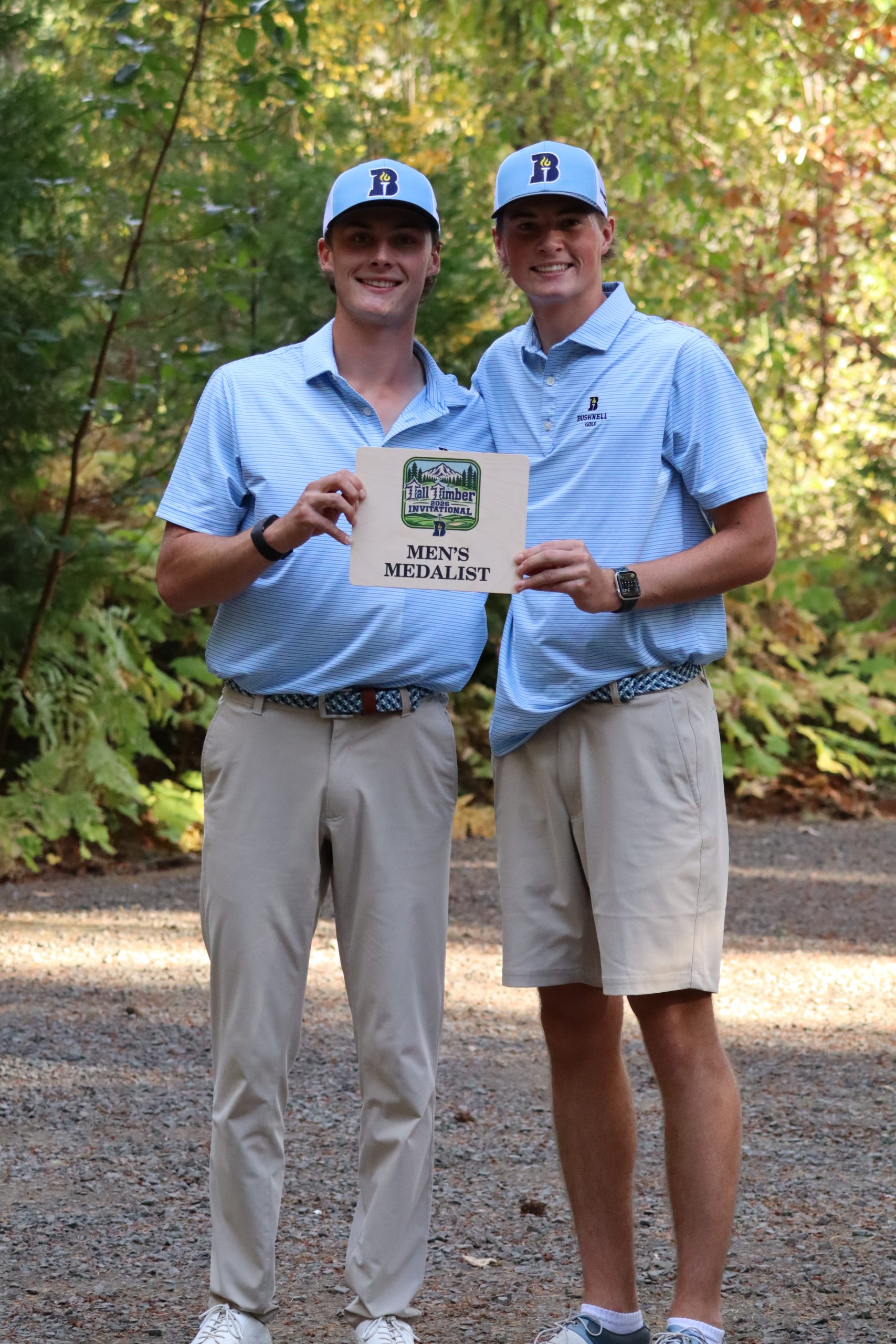 Two Bushnell University men's golfers pose in matching light blue polos and caps while holding a Tall Timber Invitational men's medalist award.