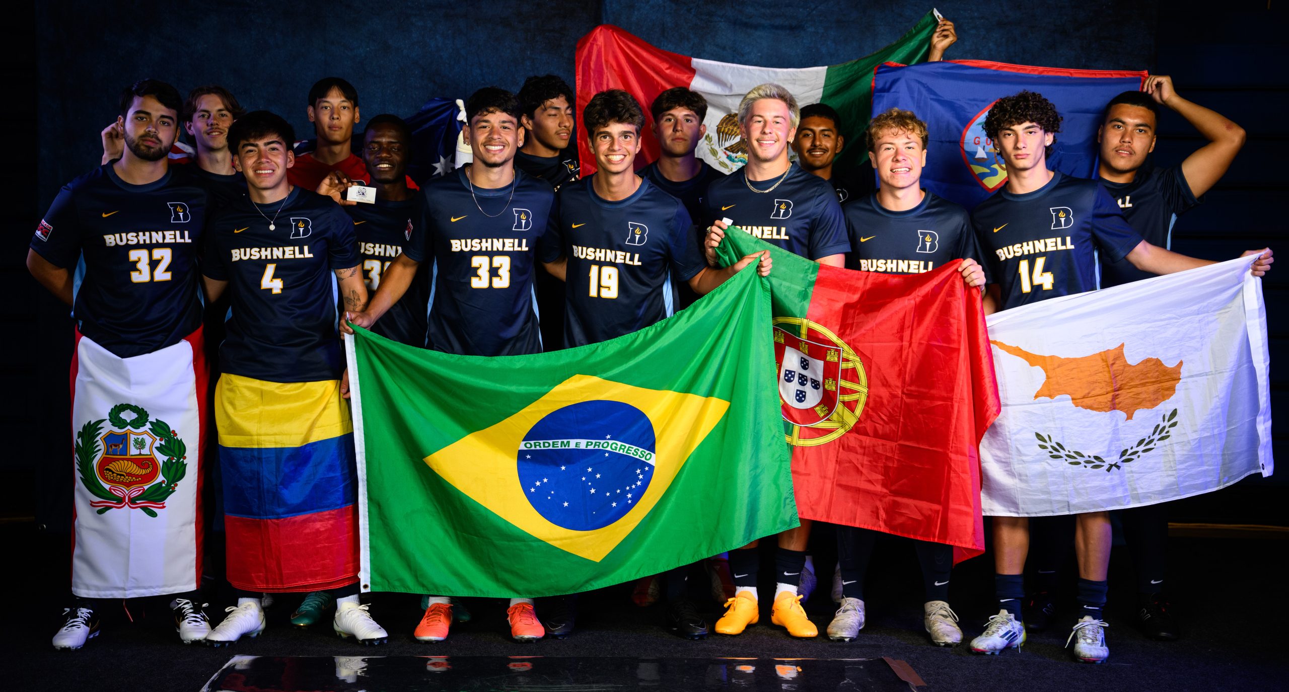 Bushnell University men's soccer players pose in navy uniforms while displaying international flags representing Peru, Colombia, Brazil, Mexico, Portugal, Cyprus and Guam.