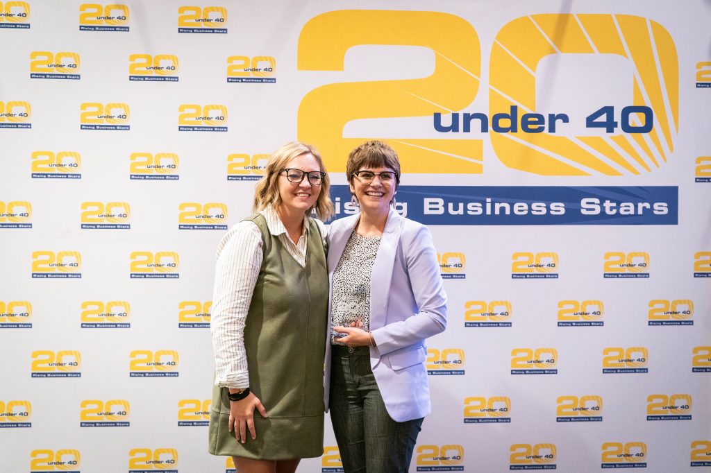 Two women smile and pose together, one holding an award, in front of the 20 Under 40 Rising Business Stars backdrop.