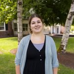 Melissa “Missy” Andersen stands outdoors on the Bushnell University campus, wearing a light blue cardigan over a black dress, with trees and a campus building in the background.