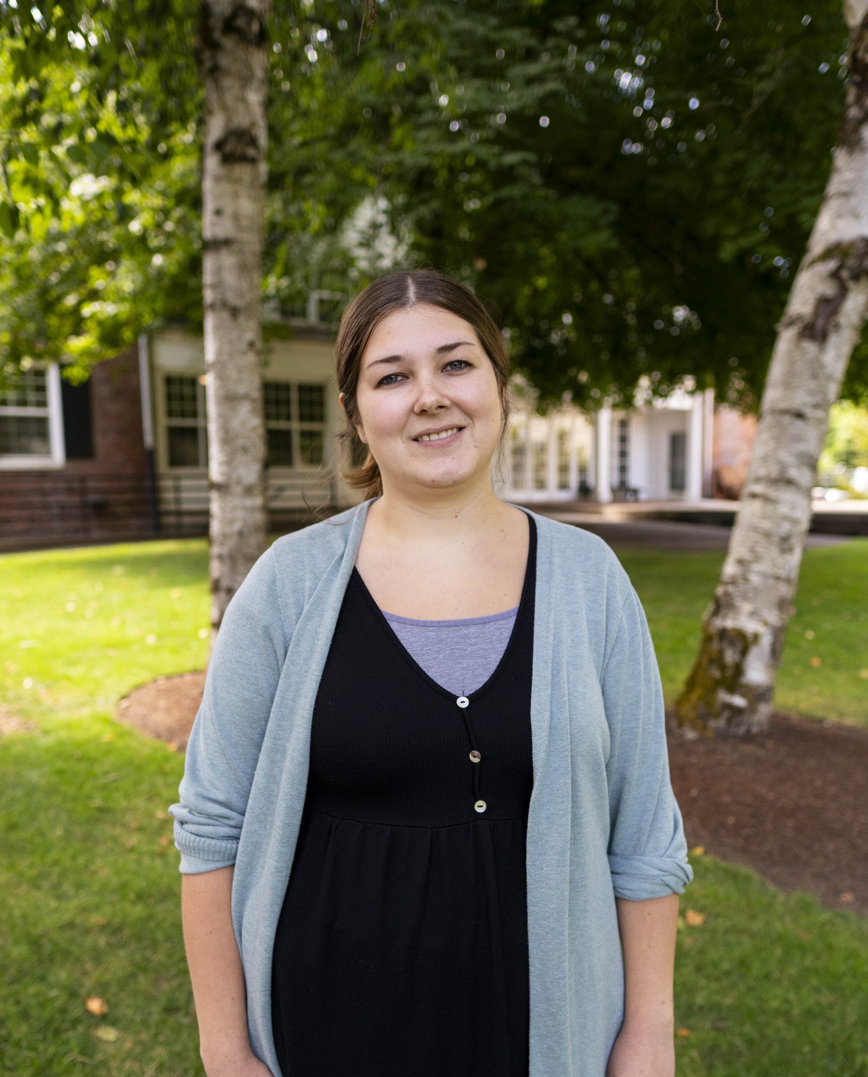 Melissa “Missy” Andersen stands outdoors on the Bushnell University campus, wearing a light blue cardigan over a black dress, with trees and a campus building in the background.