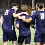 Bushnell University men's soccer players in navy uniforms gather with their arms around one another during a nighttime match.